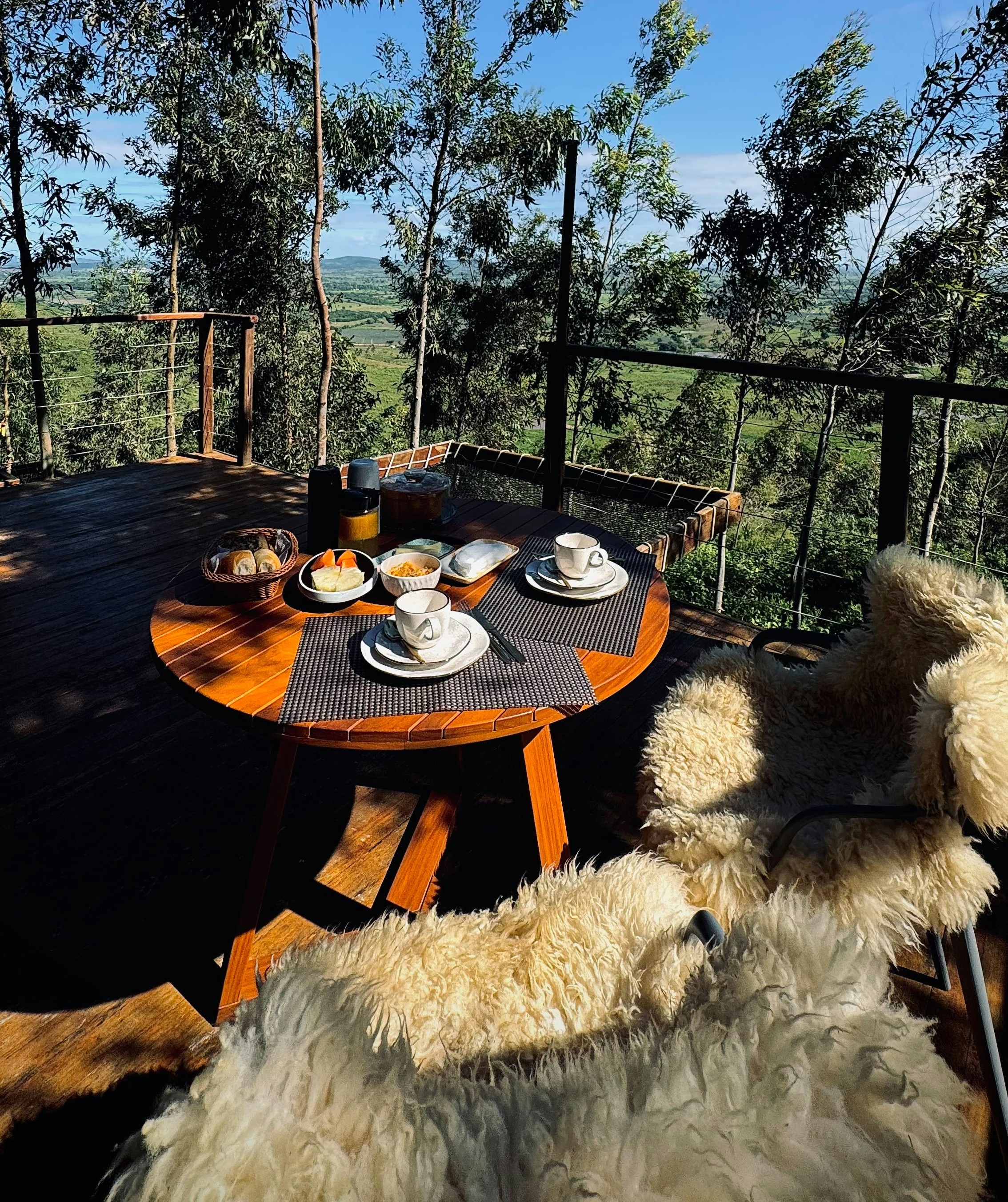 Breakfast for two on a round wooden deck table with coffee cups, fruit, and bread, chairs draped in sheepskin rugs, overlooking a green valley